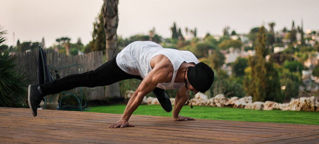 Person in a black beanie and white tank top performing a planche exercise on a wooden deck, with a garden and a cityscape in the background.